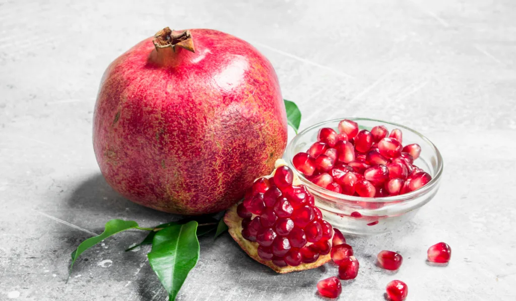 Pomegranate seeds in a bowl and a whole pomegranate with leaves.
